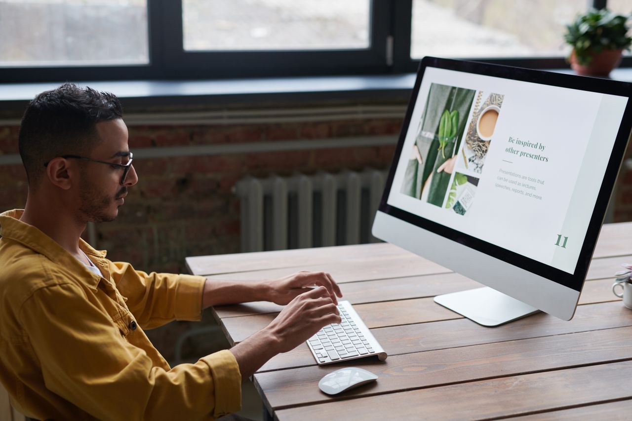 A young man in casual wear works on a computer at a wooden table in a modern, stylish office.